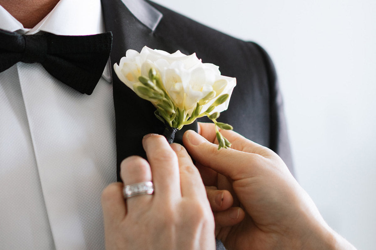 Florist Kate Hill attaching a buttonhole to the suit of a gentleman wearing a white shirt black jacket and black bow tie.