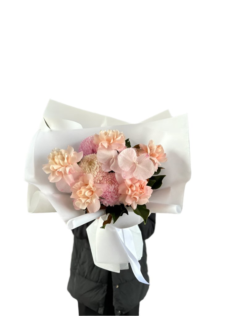 Wide image of the Chloe Blush Flower Bouquet. A close up of a blush pink flower bouquet, beautifully wrapped in white signature paper. Florist holding the bouquet in front of a white wall.