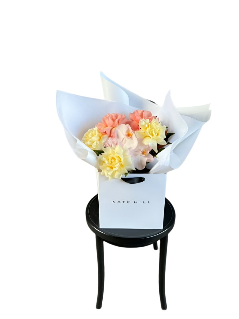 A medium sized lemon and blush guft bouquet in kate hill flower bag, sitting on a black bentwood chair in front of a white wall.