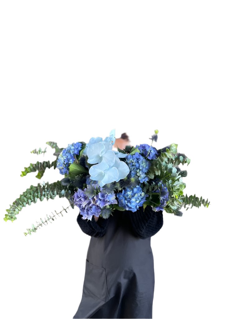 Florist holding a table hedge of blue seasonal flowers and grey blue foliages. Florist standing in front of a white wall.