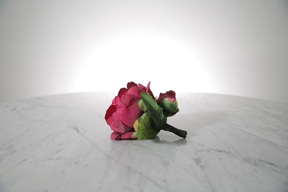 Kate Hill Flowers – artificial burgundy peony rose buttonhole flower with green leaf backing and pearl pin on marble table setting.