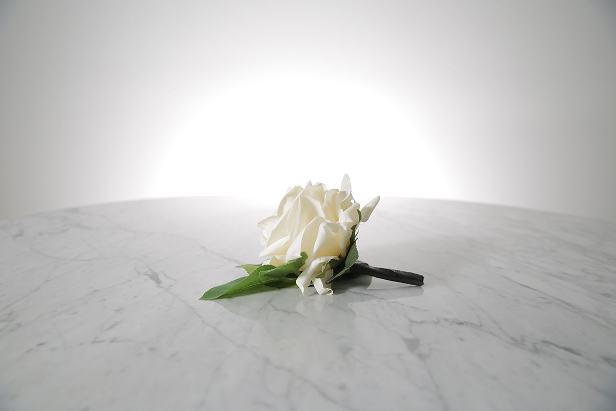 Kate Hill Flowers – artificial white rose buttonhole flower with green leaf backing, black-taped stem and pearl pin on a marble table setting.