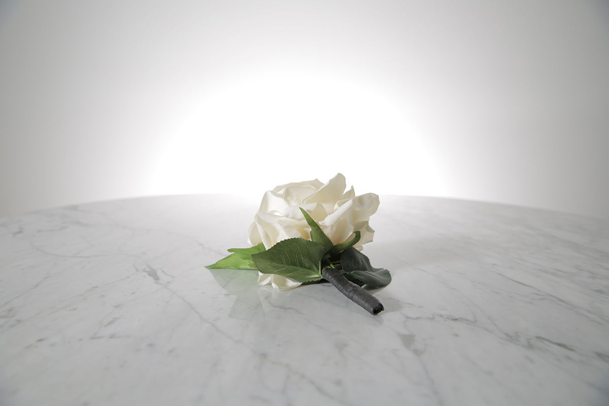 Kate Hill Flowers – artificial white rose buttonhole flower with green leaf backing, black-taped stem and pearl pin on a marble table setting.