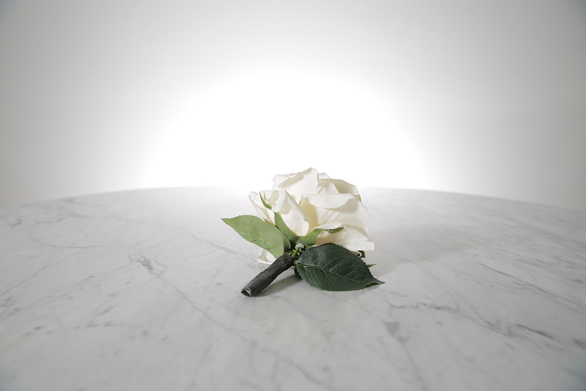 Kate Hill Flowers – artificial white rose buttonhole flower with green leaf backing, black-taped stem and pearl pin on a marble table setting.