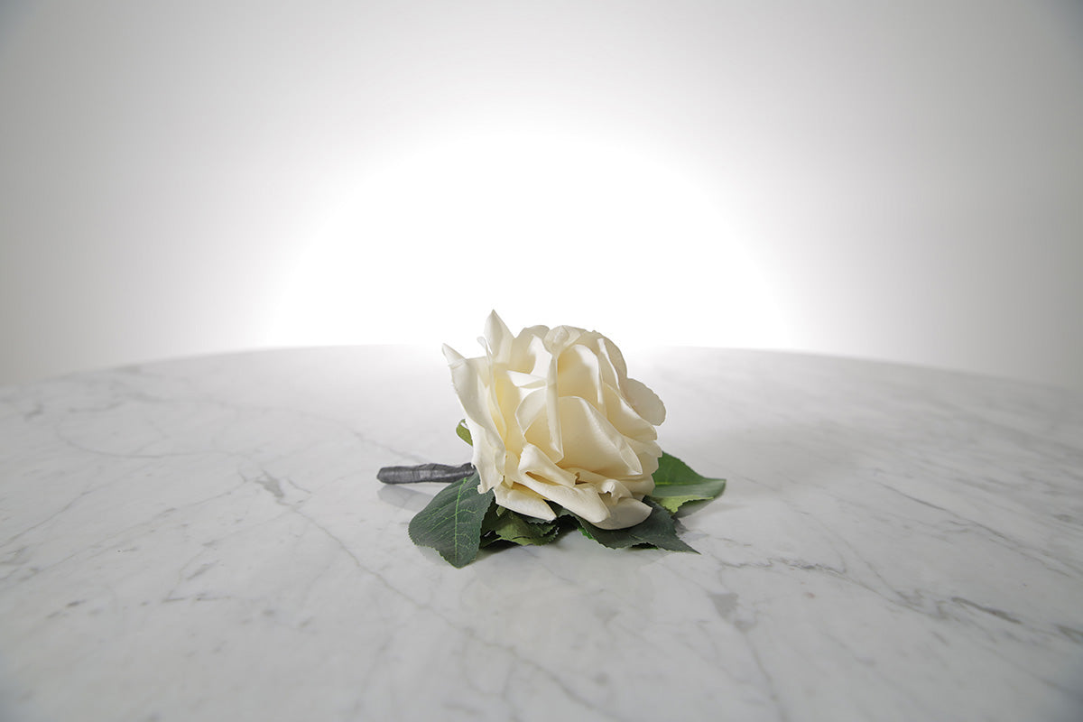 Kate Hill Flowers – artificial white rose buttonhole flower with green leaf backing, black-taped stem and pearl pin on a marble table setting.
