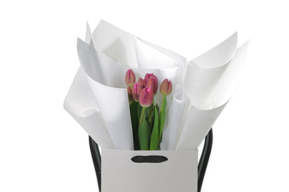 Close up image of the 10 stems of pink tulips, beautifully wrapped in signature white wrapping.  A bunch of 10 pink tulips wrapped beautifully in white signature wrapping paper and bouquet is placed into Kate Hill gift bag for presentation. Bag of pink tulips sitting on a black bentwood chair for photograph. Bouquet stands approximately 45cm tall on chair.