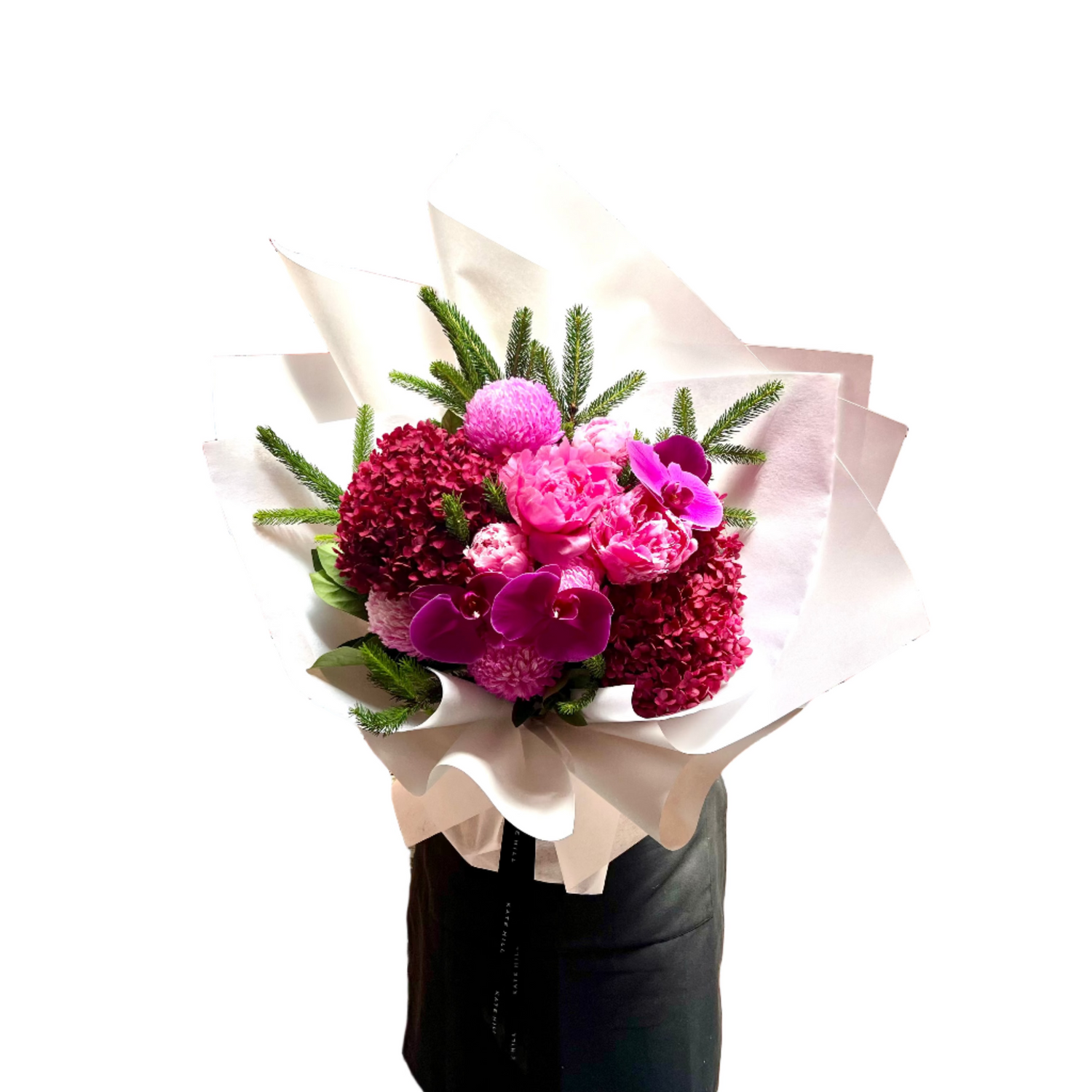 A large bouquet of flowers in hot pink, magenta and burgundy colours, detailed with a hint of Christmas spruce foliage. Bouquet is wrapped beautifully in white signature wrapping and florist is standing against a white wall.