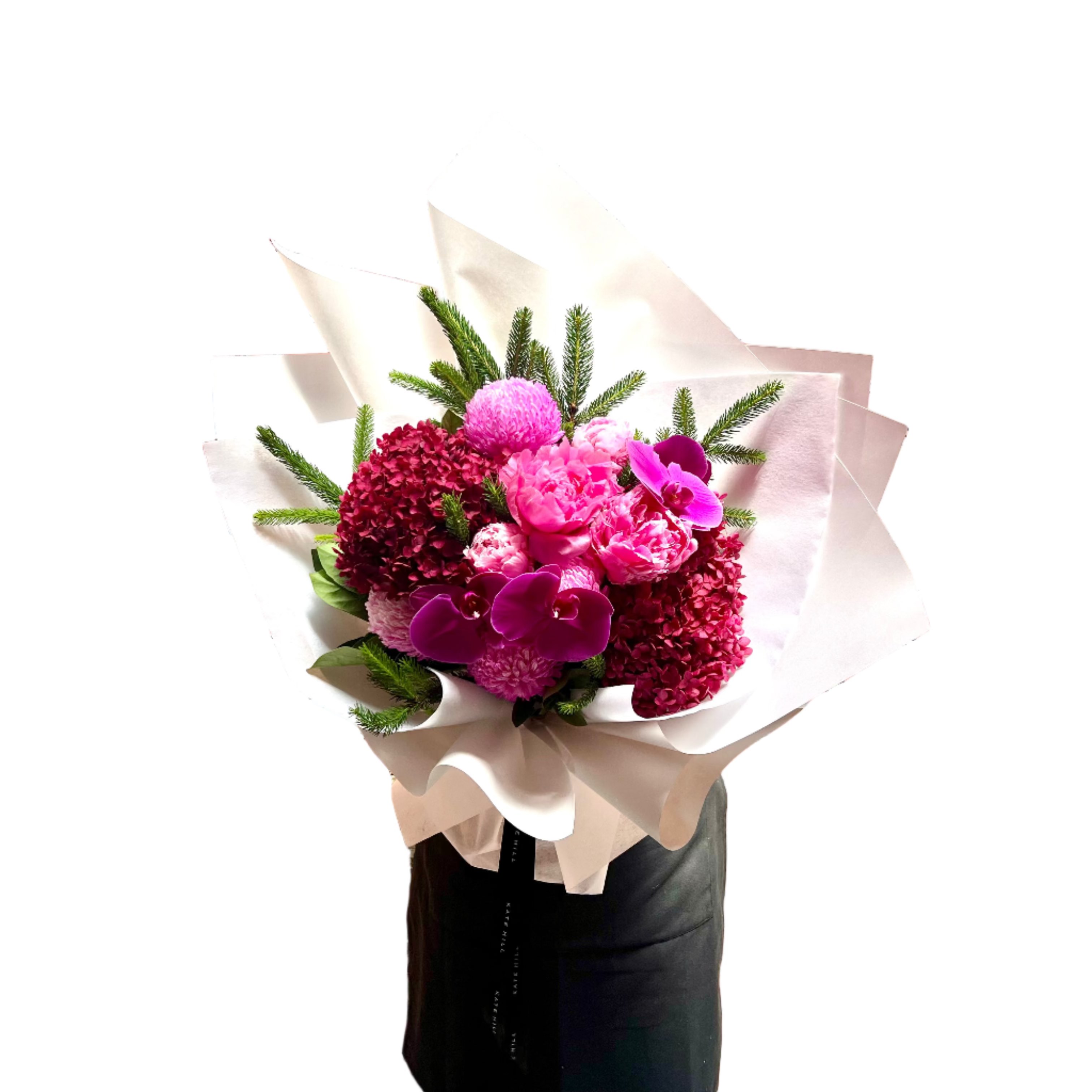 A large bouquet of flowers in hot pink, magenta and burgundy colours, detailed with a hint of Christmas spruce foliage. Bouquet is wrapped beautifully in white signature wrapping and florist is standing against a white wall.