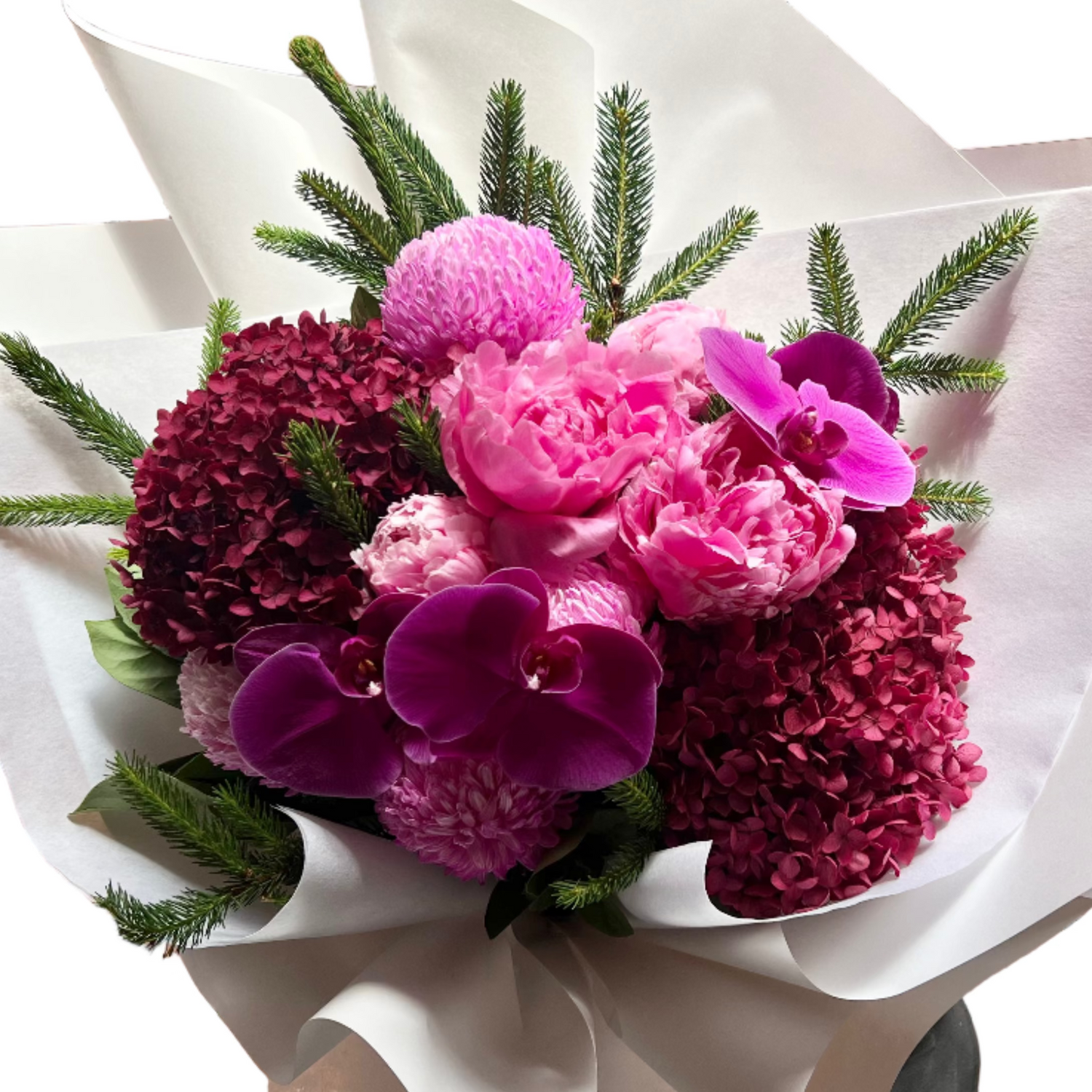 Wide image of a large bouquet of flowers in hot pink, magenta and burgundy colours, detailed with a hint of Christmas spruce foliage. Bouquet is wrapped beautifully in white signature wrapping and florist is standing against a white wall.