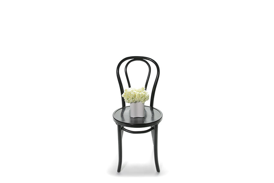 Wide image of Earl wave vase. A miniature white wave ceramic vase, displaying a dense dome of erlicheer flowers. Small wave vase sitting on black bentwood chair with white background.