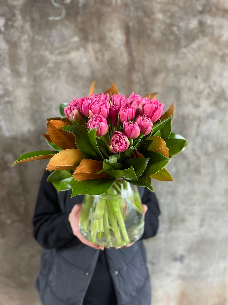 Florist holding a clear tapered vase full of pink double tulips and magnolia foliage, in front of a concrete wall.