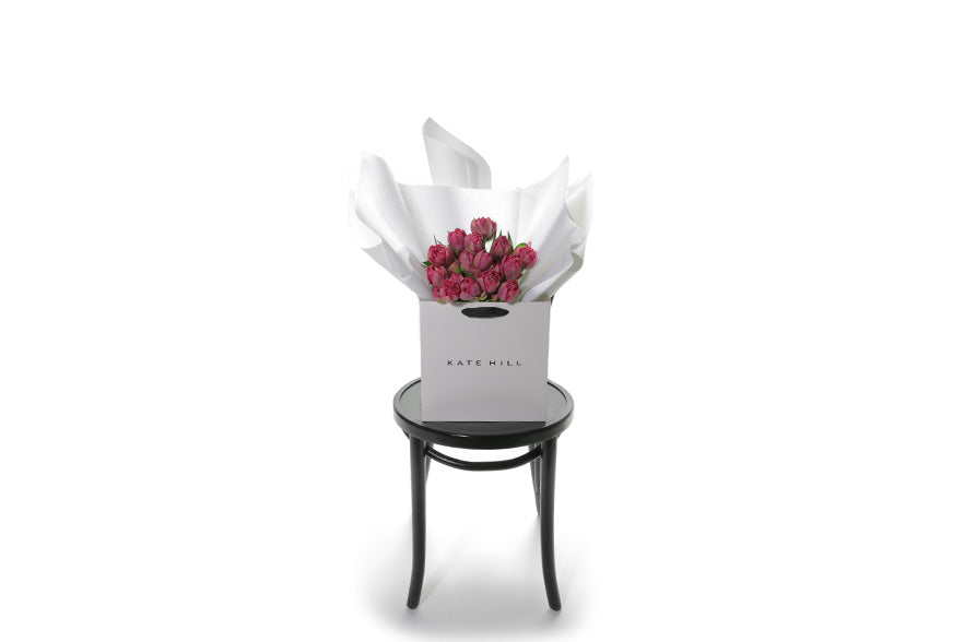 Wide image of FRANKIE double pink tulip bouquet. A wrapped bouquet of 20 stems of dark double pink tulips. FRANKIE double Tulip bouquet is beautifully gift wrapped in Kate Hill Flowers signature style that includes layers of premium white and silk paper, finished with Kate Hill signature ribbon. Bouquet is placed into our Kate Hill flower bag. Bouquet bag is sitting on a black bentwood chair. Image gives a honest idea of the bouquet size.