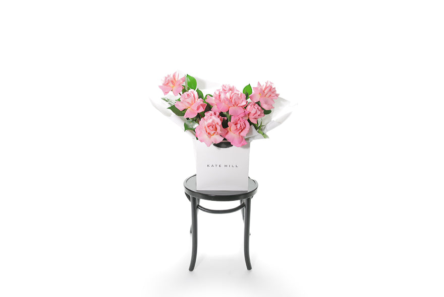 HERMOSA Rose Bouquet is our best selling watermelon pink rose bouquet, curated with Reflexed watermelon pink roses and green seasonal foliage. Large bouquet of watermelon roses presented in our luxury flower bag so it stays fresh in water. Bouquet bag sitting on a black bentwood chair. Wide image to show the true size of the bouquet.