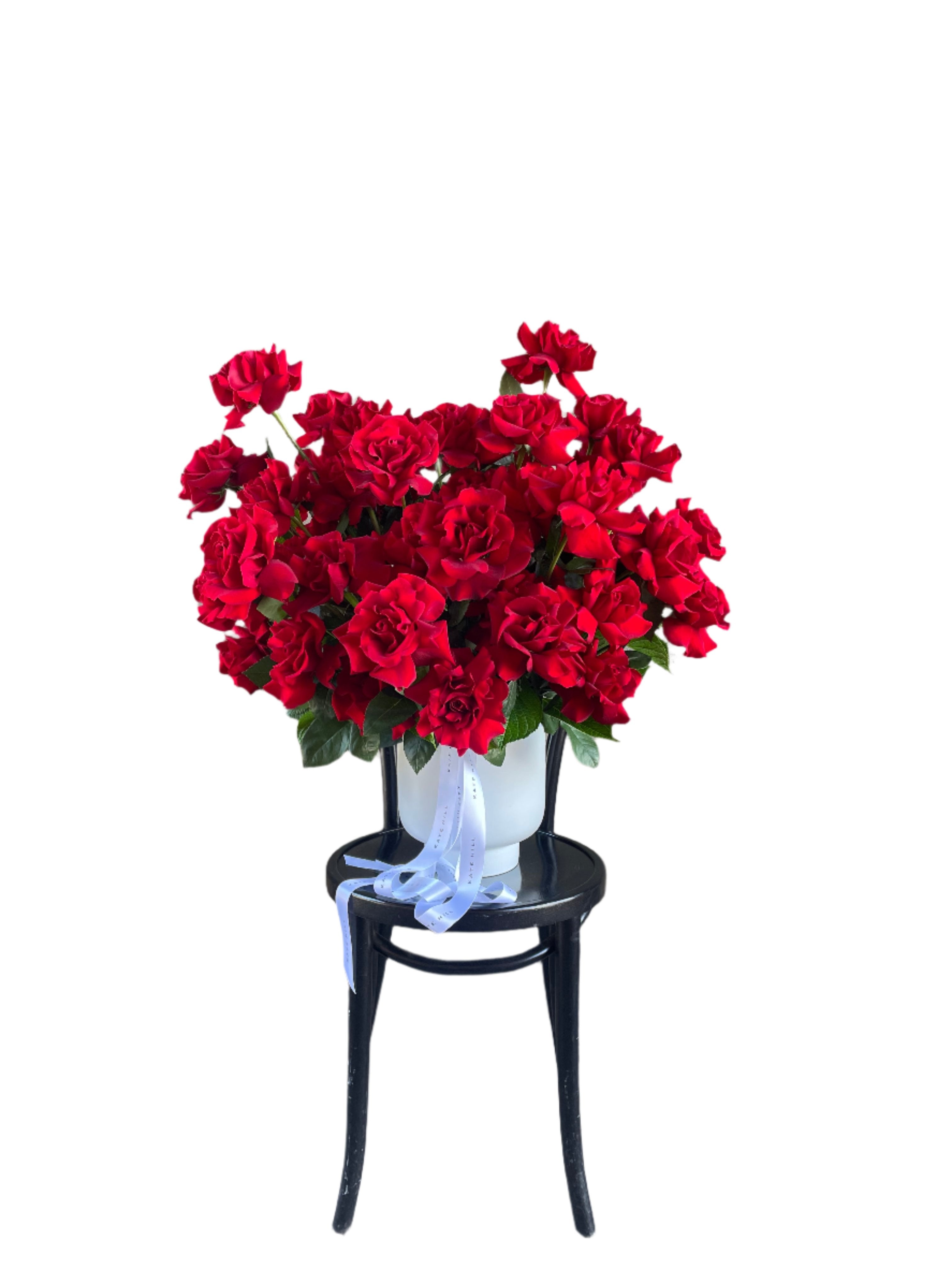 A grande design, displaying 50 long stemmed red roses on mass in a large white footed ceramic vase. Large design is sitting on a black bentwood chair with white wall in the background.