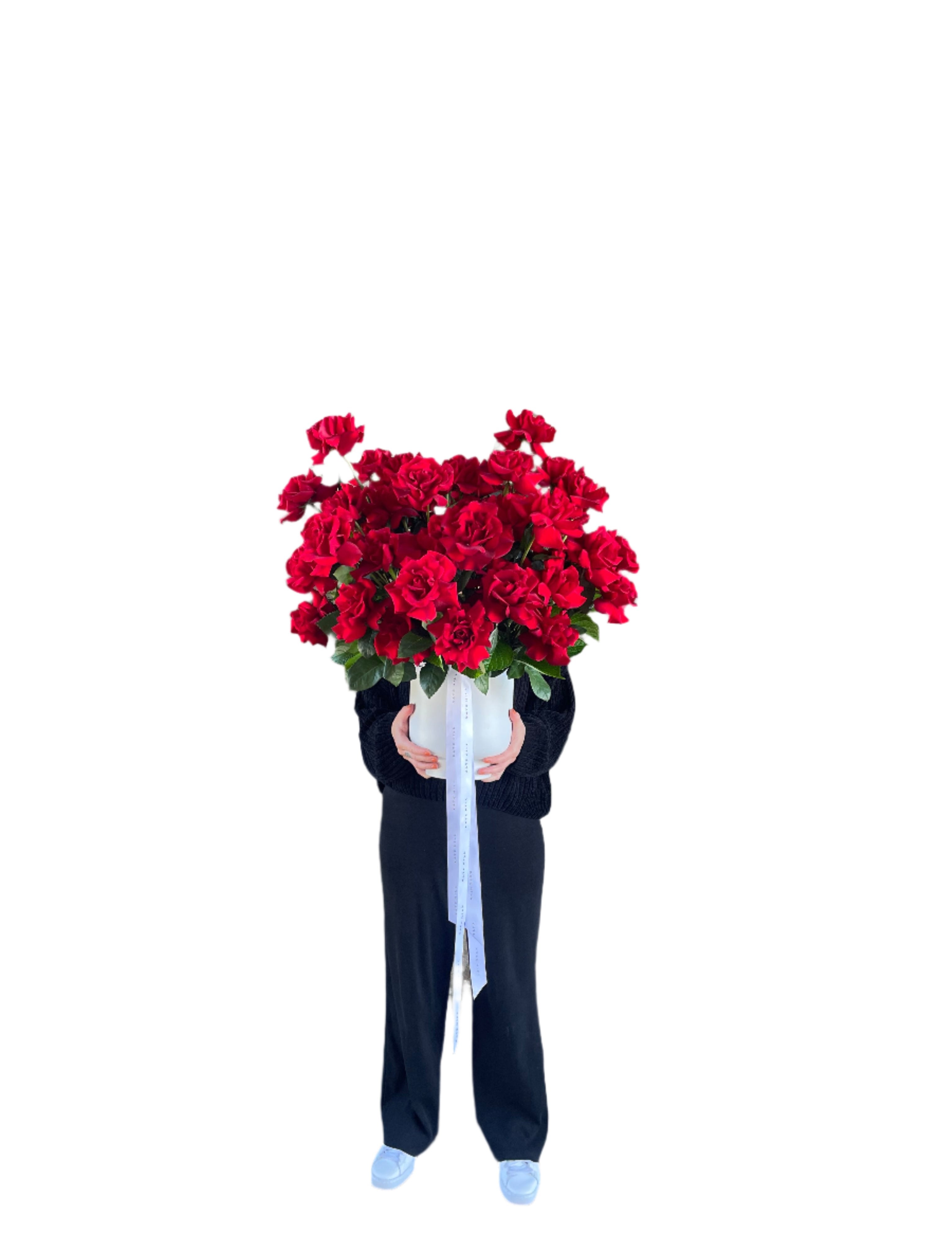 Florist holding in full view, A grande design, displaying 50 long stemmed red roses on mass in a large white footed ceramic vase.