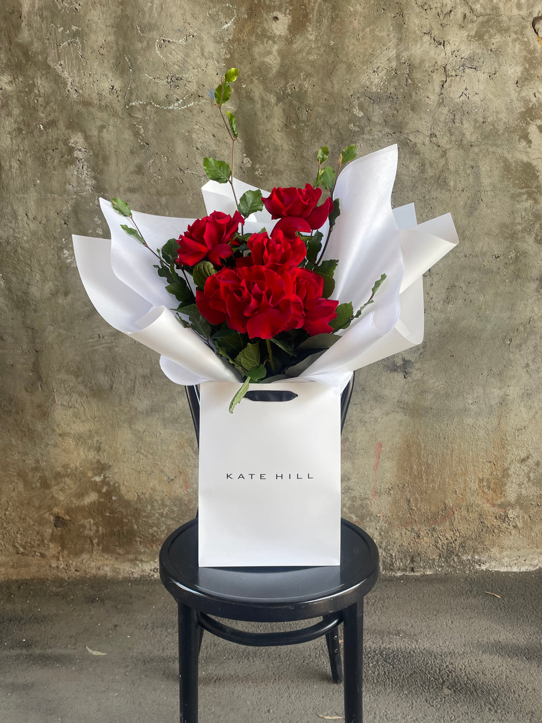 Simple red rose bouquet displaying 6 stems of red reflexed roses. Valentines day or Romance gift bouquet presented in our Kate Hill Flower Bag. Red rose bouquet bag sitting on a black bentwood chair with white background.
