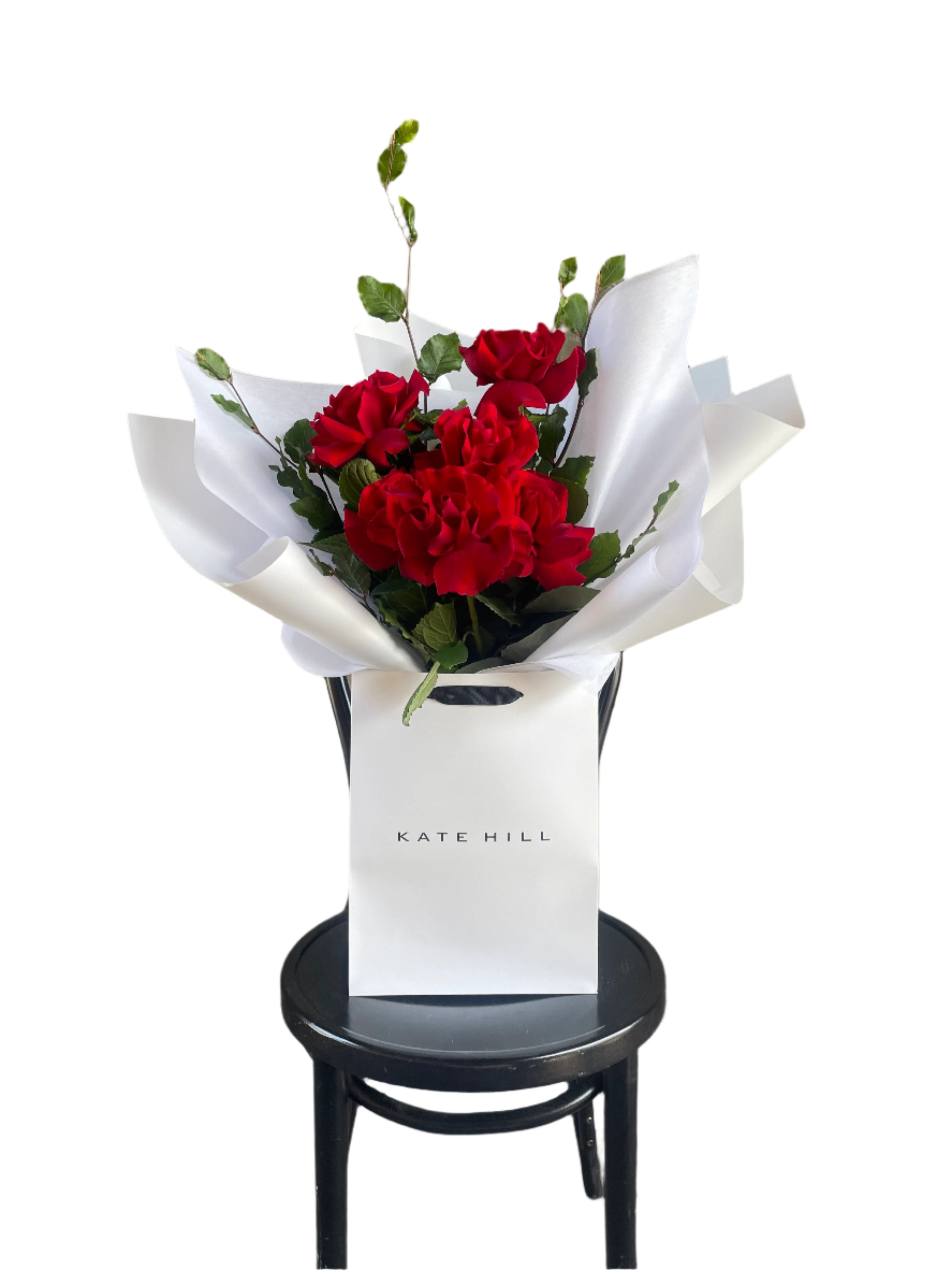 Simple red rose bouquet displaying 6 stems of red reflexed roses. Valentines day or Romance gift bouquet presented in our Kate Hill Flower Bag. Red rose bouquet bag sitting on a black bentwood chair with white background.