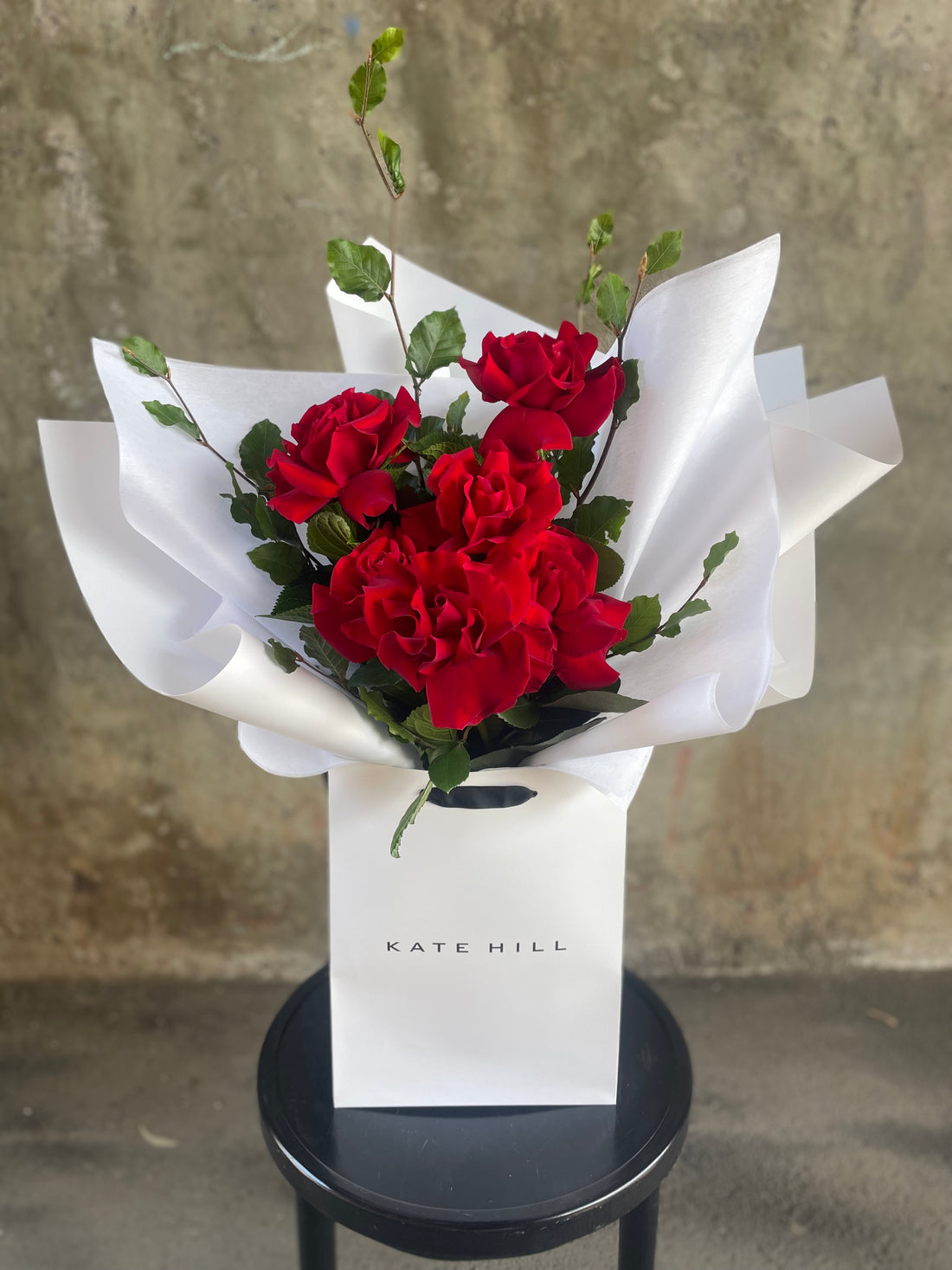 Close up image of the red rose bouquet. Simple red rose bouquet displaying 6 stems of red reflexed roses. Valentines day or Romance gift bouquet presented in our Kate Hill Flower Bag. Red rose bouquet bag sitting on a black bentwood chair with white background.