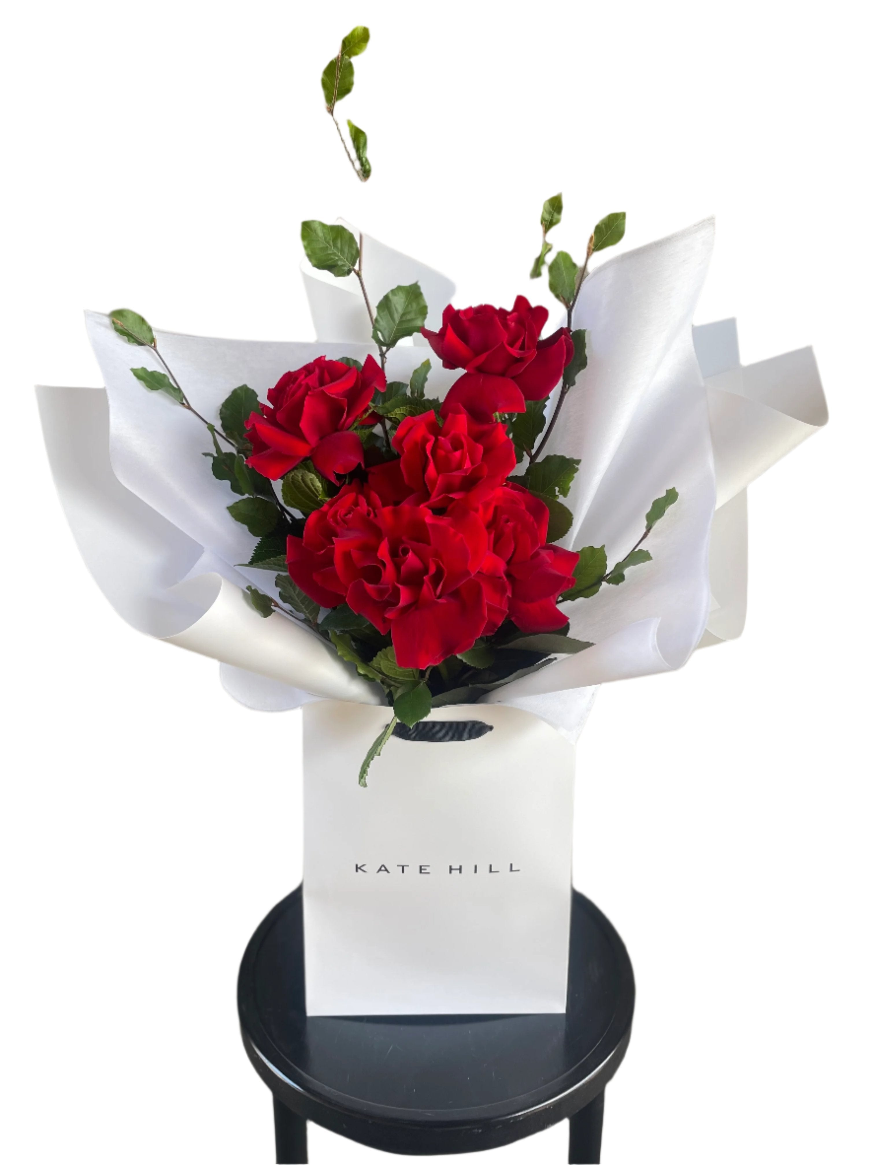 Close up image of the red rose bouquet. Simple red rose bouquet displaying 6 stems of red reflexed roses. Valentines day or Romance gift bouquet presented in our Kate Hill Flower Bag. Red rose bouquet bag sitting on a black bentwood chair with white background.