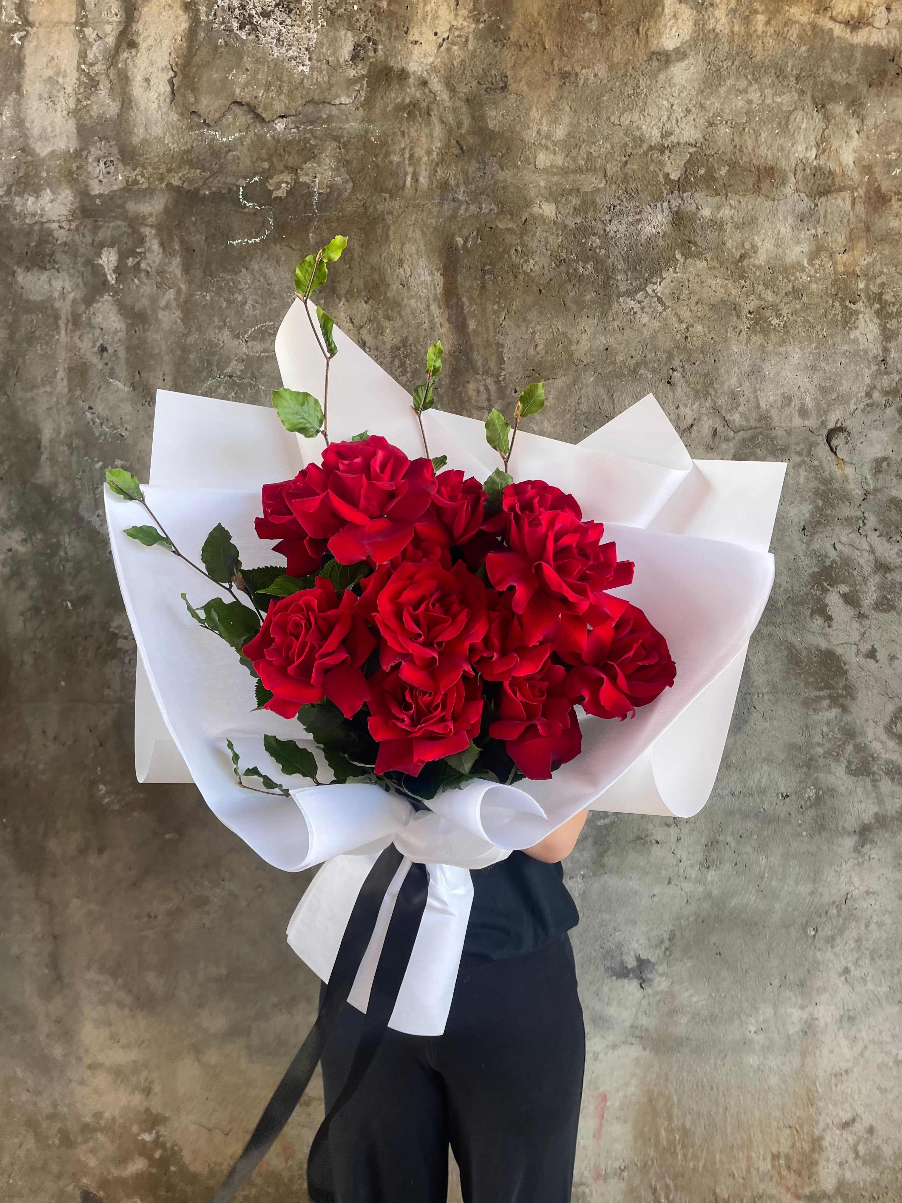 Bouquet of one dozen red roses and foliage. A luxe red rose bouquet designed with 12 premium red roses and foliages, beautifully wrapped with signature KHF wrapping. Florist holding the bouquet in front of a concrete wall.