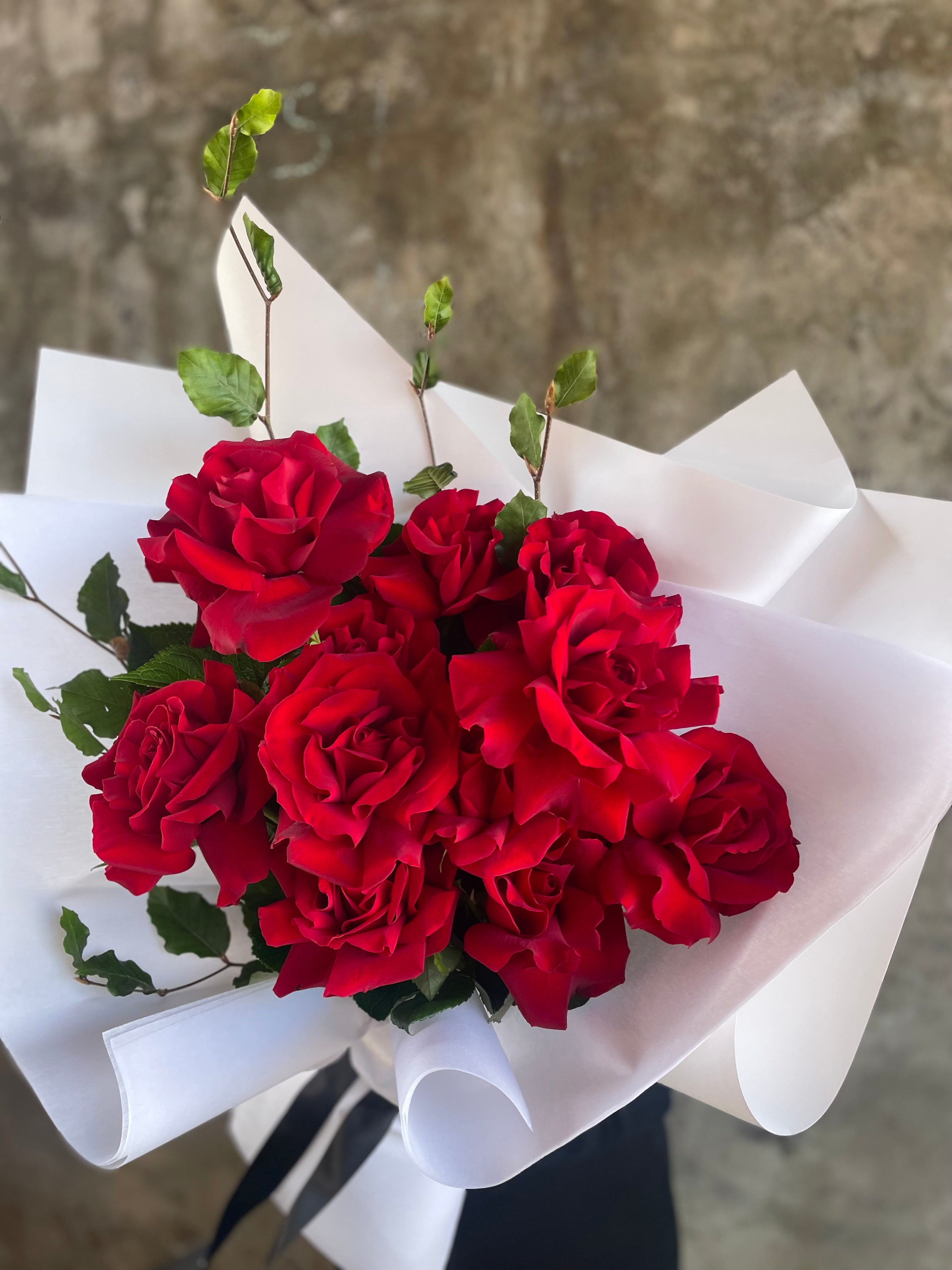 Close up image of 1 dozen red rose bouquet, wrapped in white signature style. Florist holding bouquet in front of concrete wall.