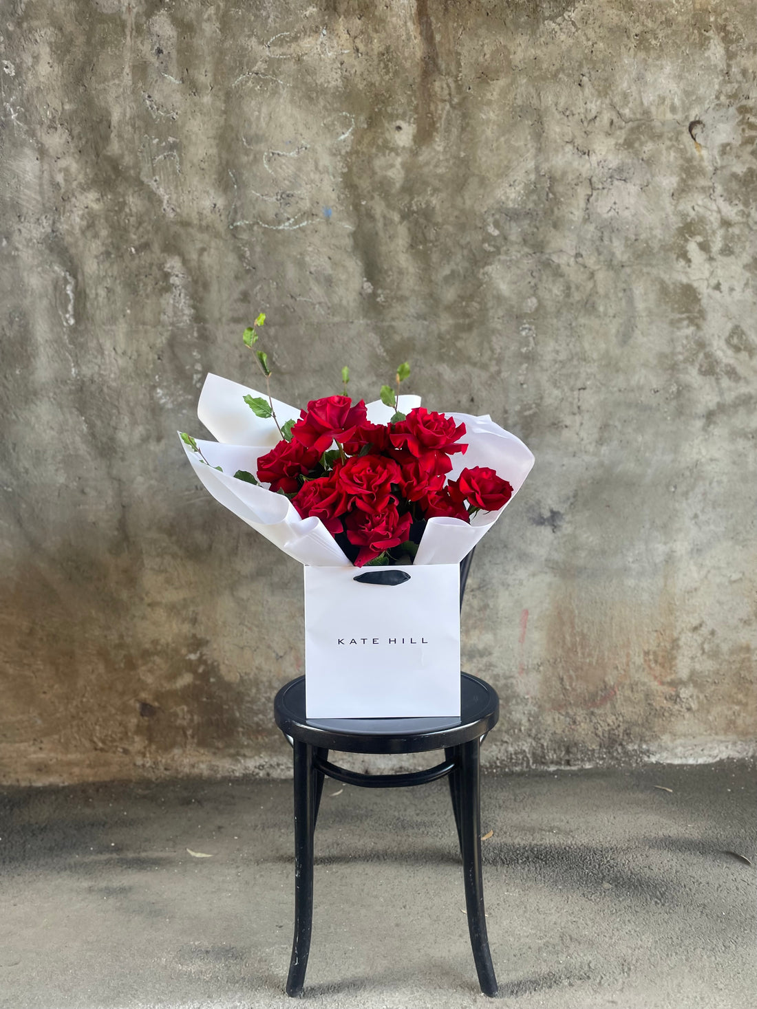 A luxe red rose bouquet designed with 12 premium red roses and foliages, beautifully wrapped with signature KHF wrapping. Bouquet displayed in KHF flower bag and sitting on a black bentwood chair with concrete wall in the background.