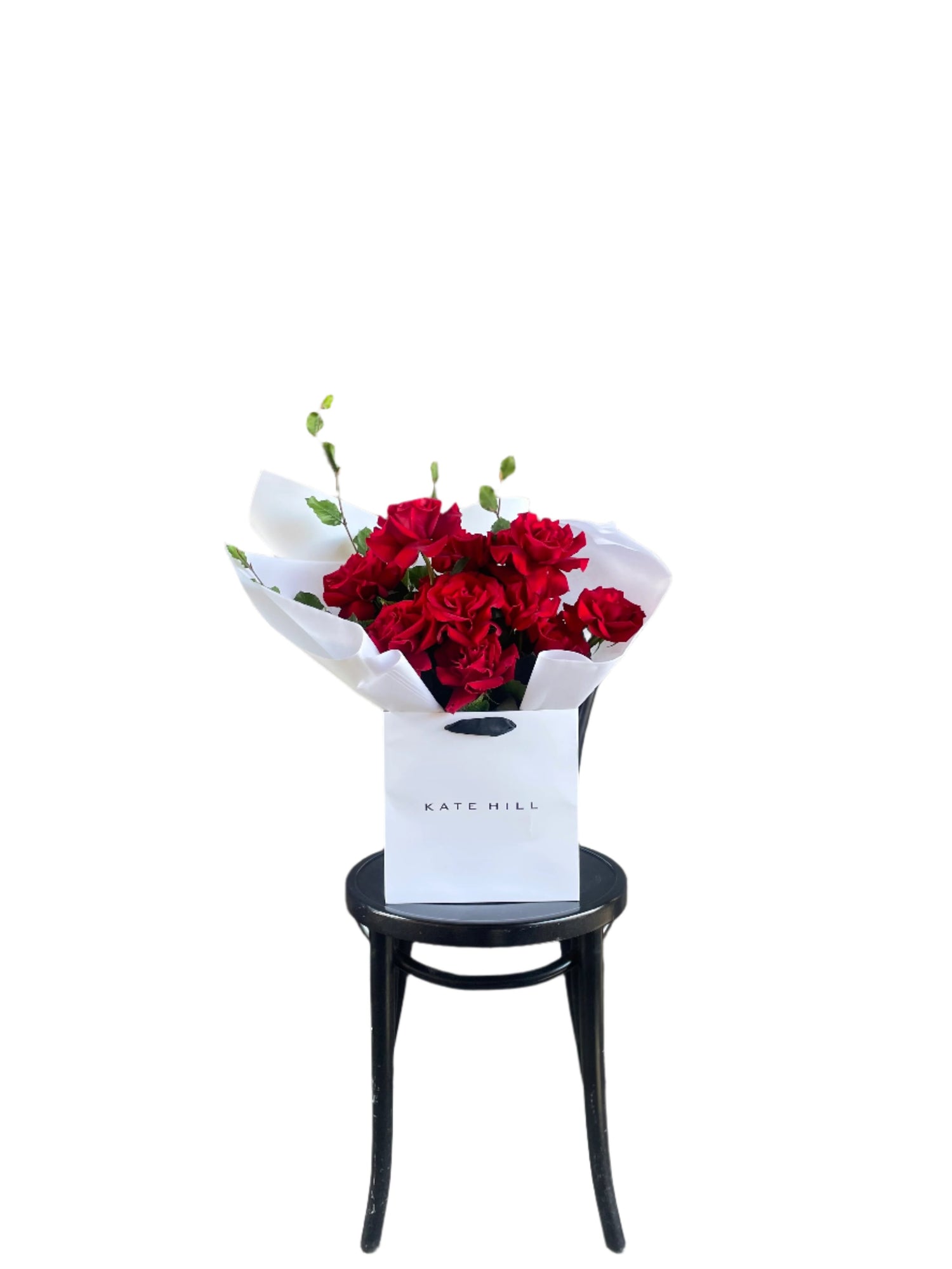 A luxe red rose bouquet designed with 12 premium red roses and foliages, beautifully wrapped with signature KHF wrapping. Bouquet displayed in KHF flower bag and sitting on a black bentwood chair with white wall in the background.
