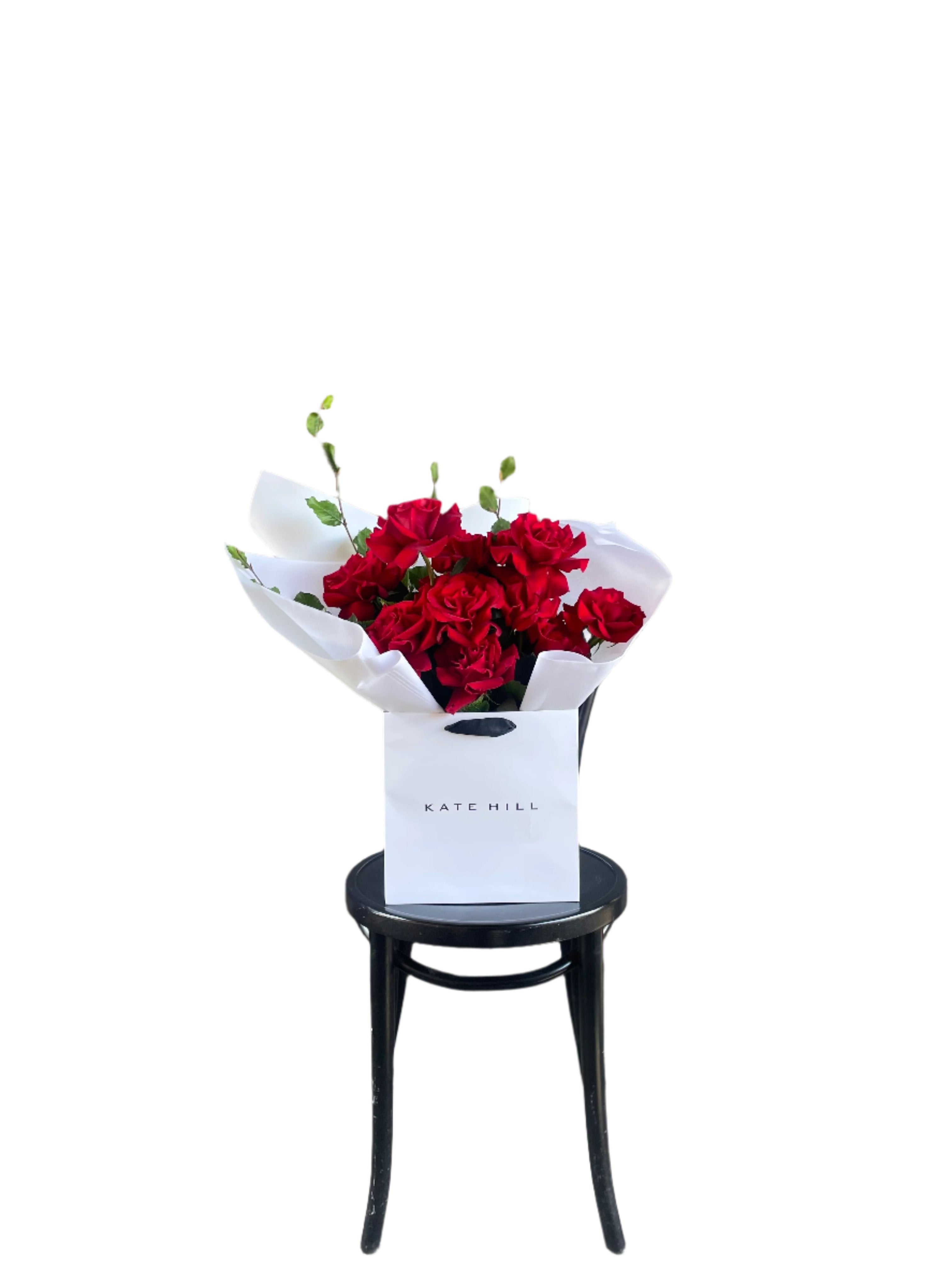 A luxe red rose bouquet designed with 12 premium red roses and foliages, beautifully wrapped with signature KHF wrapping. Bouquet displayed in KHF flower bag and sitting on a black bentwood chair with white wall in the background.