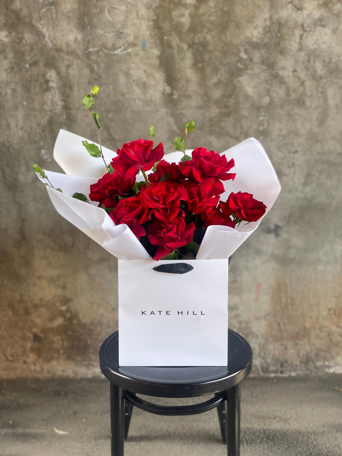 Close up image of a dozen red rose and foliage bouquet. A luxe red rose bouquet designed with 12 premium red roses and foliages, beautifully wrapped with signature KHF wrapping. Bouquet displayed in KHF flower bag and sitting on a black bentwood chair with concrete wall in the background.