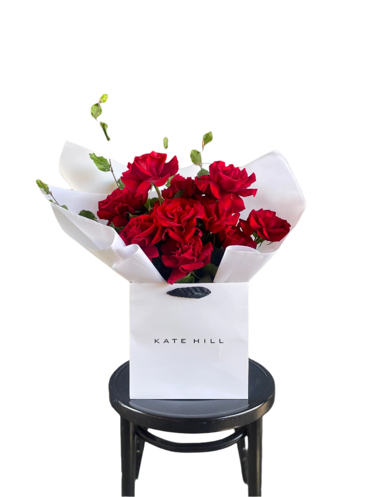 Close up image of a dozen red rose and foliage bouquet. A luxe red rose bouquet designed with 12 premium red roses and foliages, beautifully wrapped with signature KHF wrapping. Bouquet displayed in KHF flower bag and sitting on a black bentwood chair with white wall in the background.
