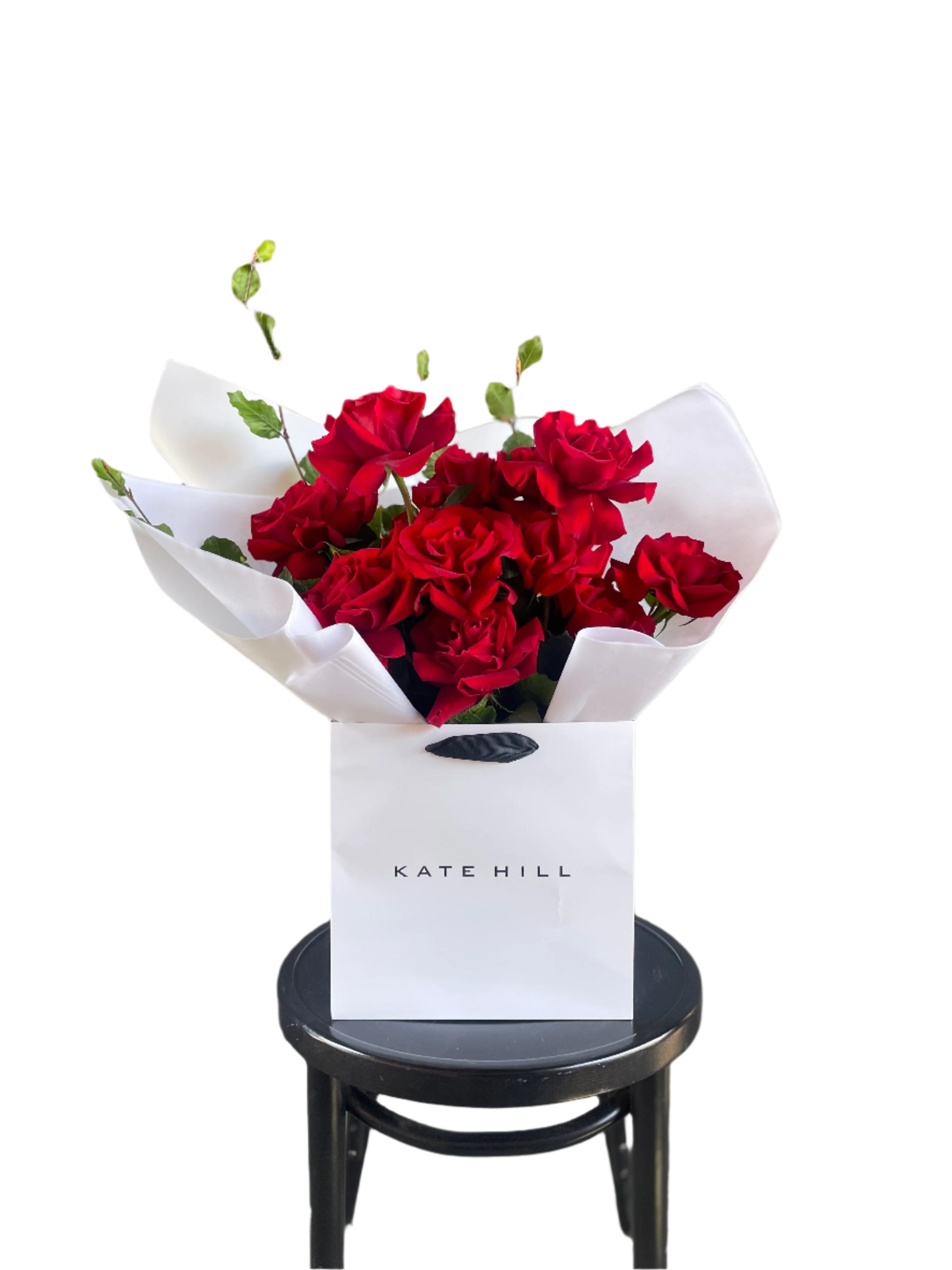 Close up image of a dozen red rose and foliage bouquet. A luxe red rose bouquet designed with 12 premium red roses and foliages, beautifully wrapped with signature KHF wrapping. Bouquet displayed in KHF flower bag and sitting on a black bentwood chair with white wall in the background.