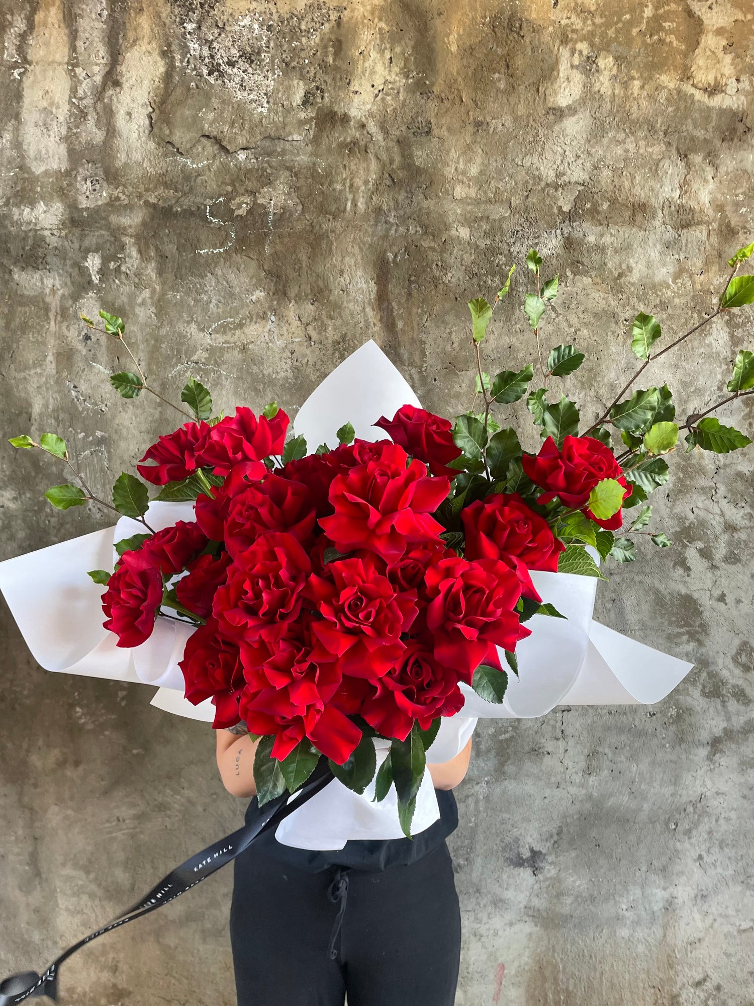Florist holding bouquet of 24 red roses and foliage. Wrapped beautifully in white signature wrapping style. Florist holding bouquet in front of a concrete wall.