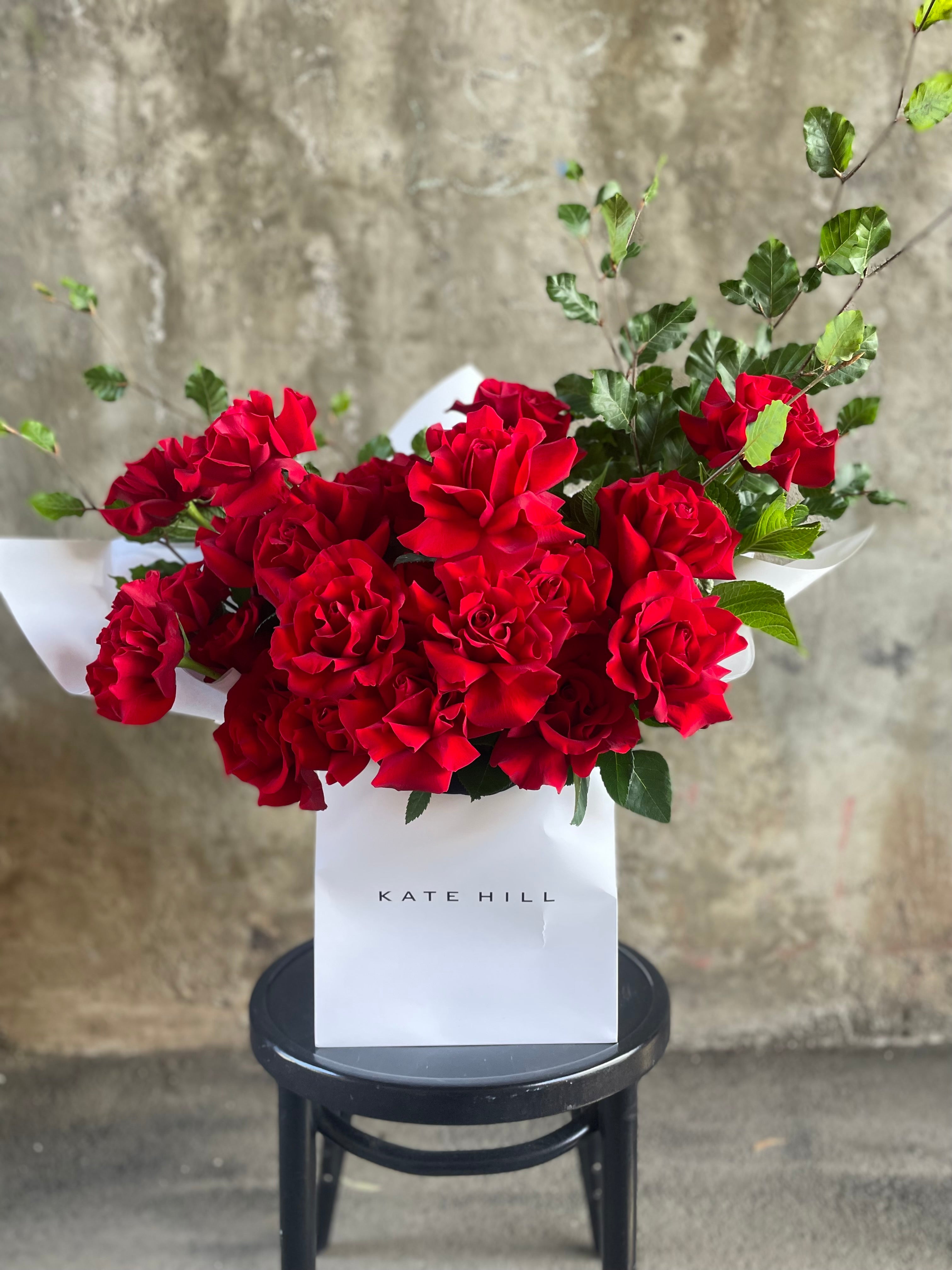 Close up image of 24 red rose bouquet in flower bag. A large red bouquet featuring 24 stems and foliage, wrapped in signature white wrapping and placed into a KHF flower bag. Bouquet bag sitting on a black bentwood chair with concrete wall in background.