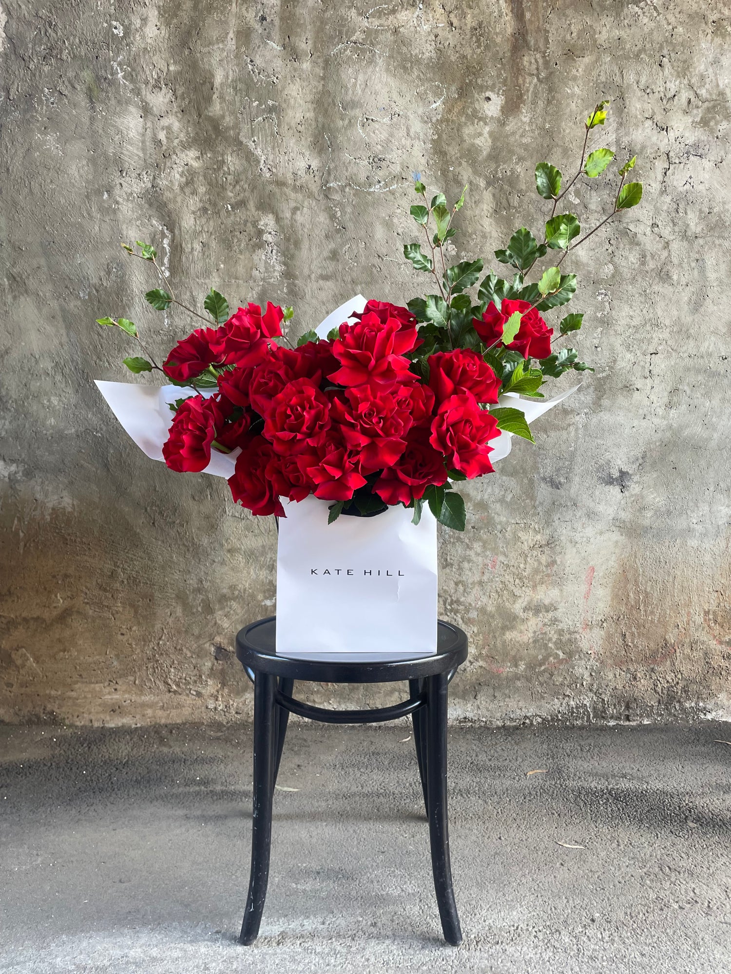 A large red bouquet featuring 24 stems and foliage, wrapped in signature white wrapping and placed into a KHF flower bag. Bouquet bag sitting on a black bentwood chair with concrete wall in background.