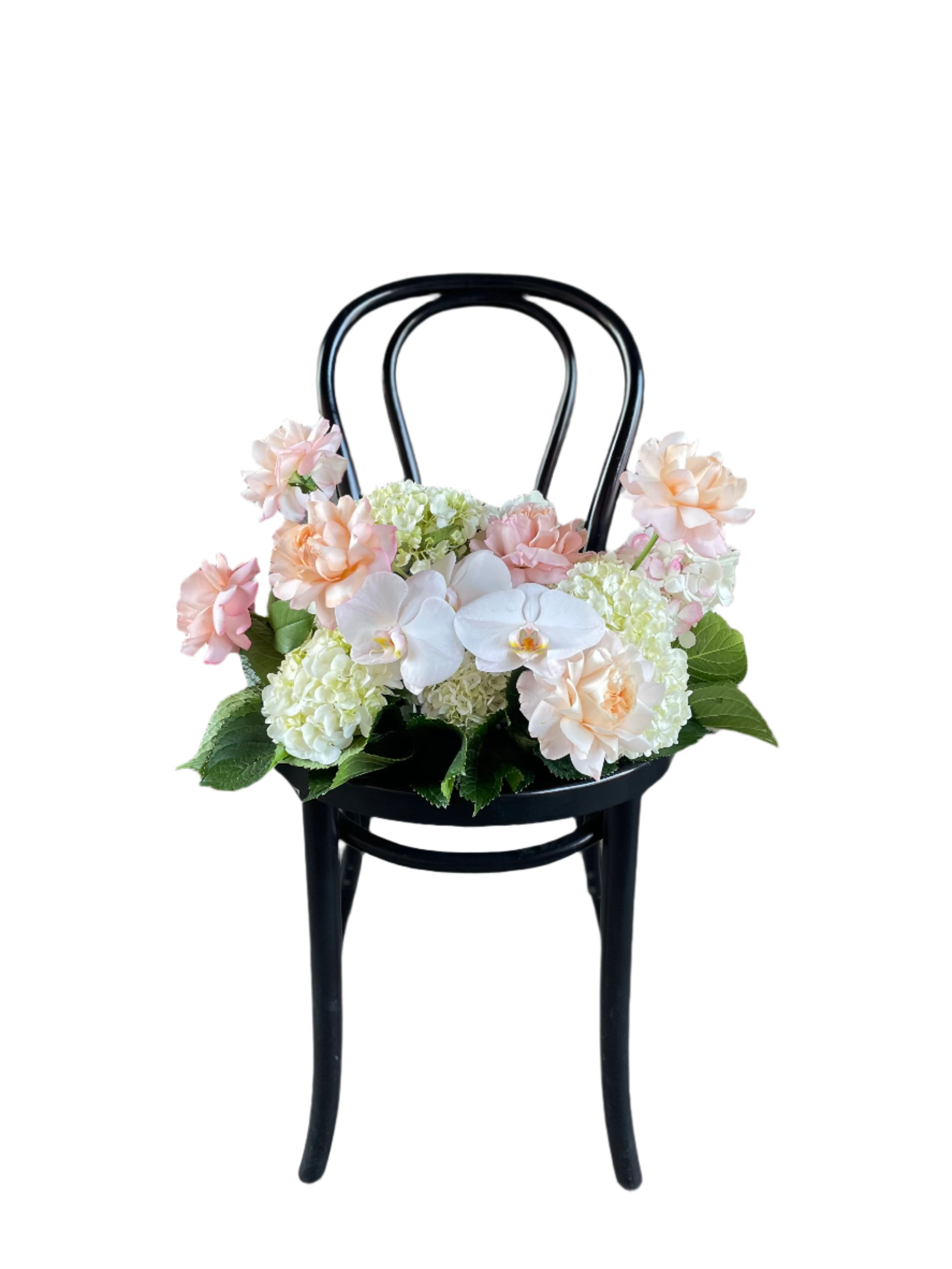 A floral hedge in white, blush and green tones, sitting on a black bentwood chair with a white wall background.