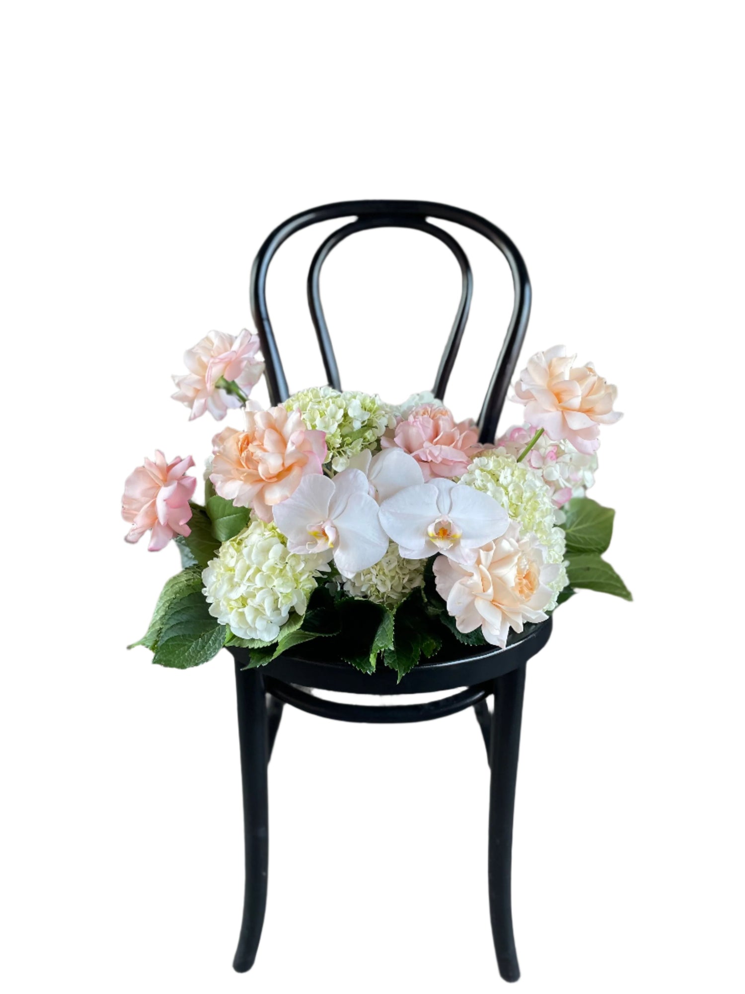 A floral hedge in white, blush and green tones, sitting on a black bentwood chair with a white wall background.