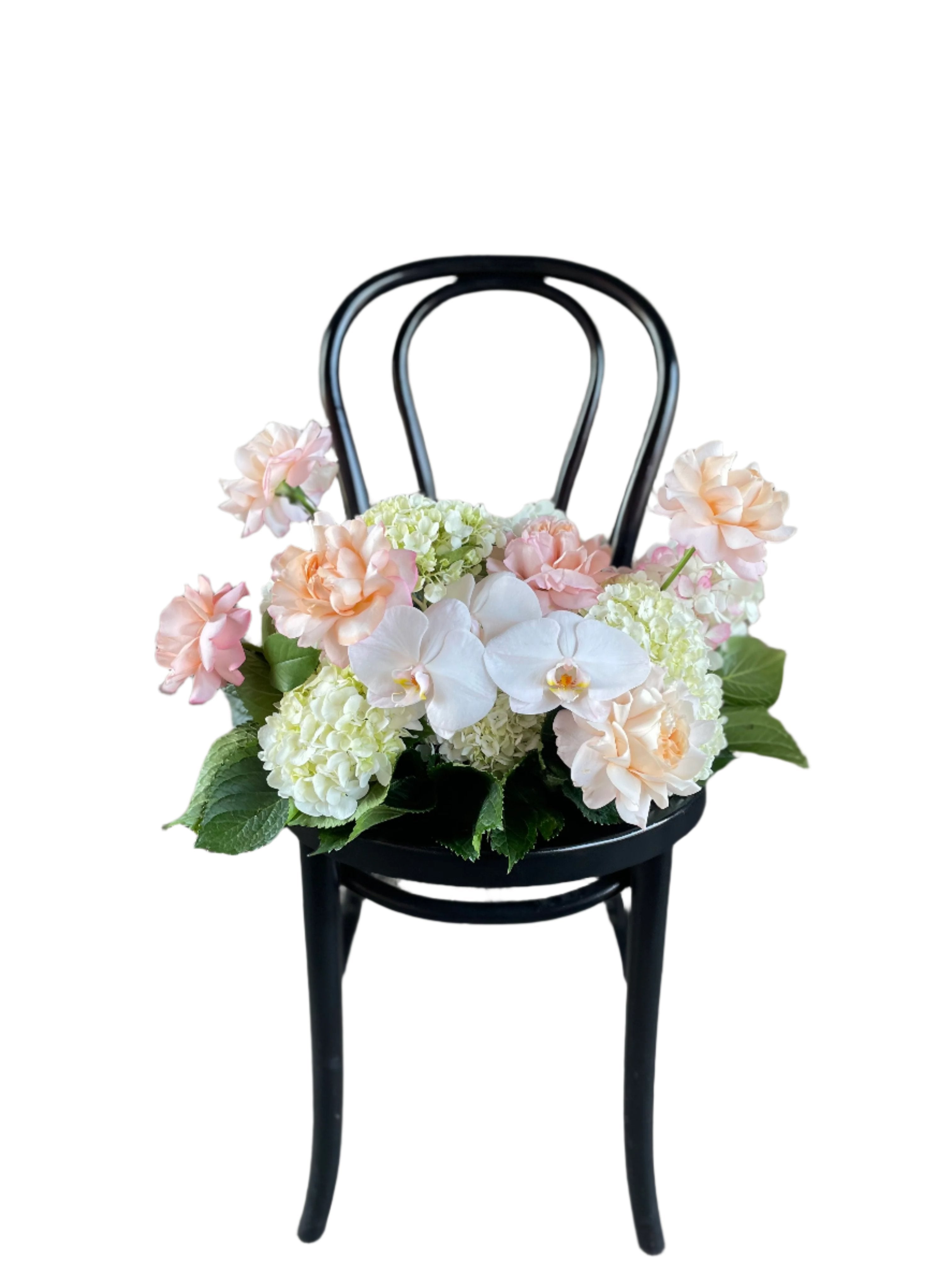 A floral hedge in white, blush and green tones, sitting on a black bentwood chair with a white wall background.