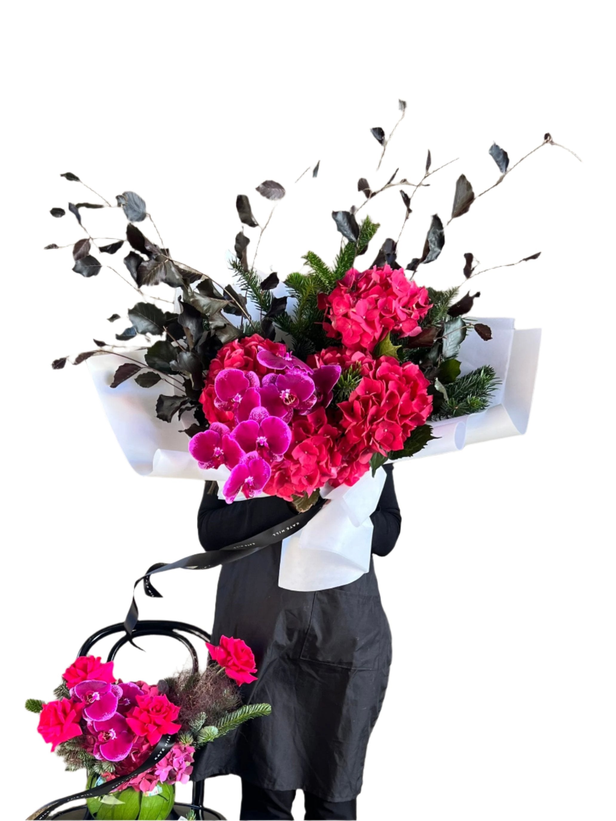 Florist holding a raspberry, burgundy and magenta Christmas flower bouquet and a complimentary vase design sitting on a lack bentwood chair with white wall background.