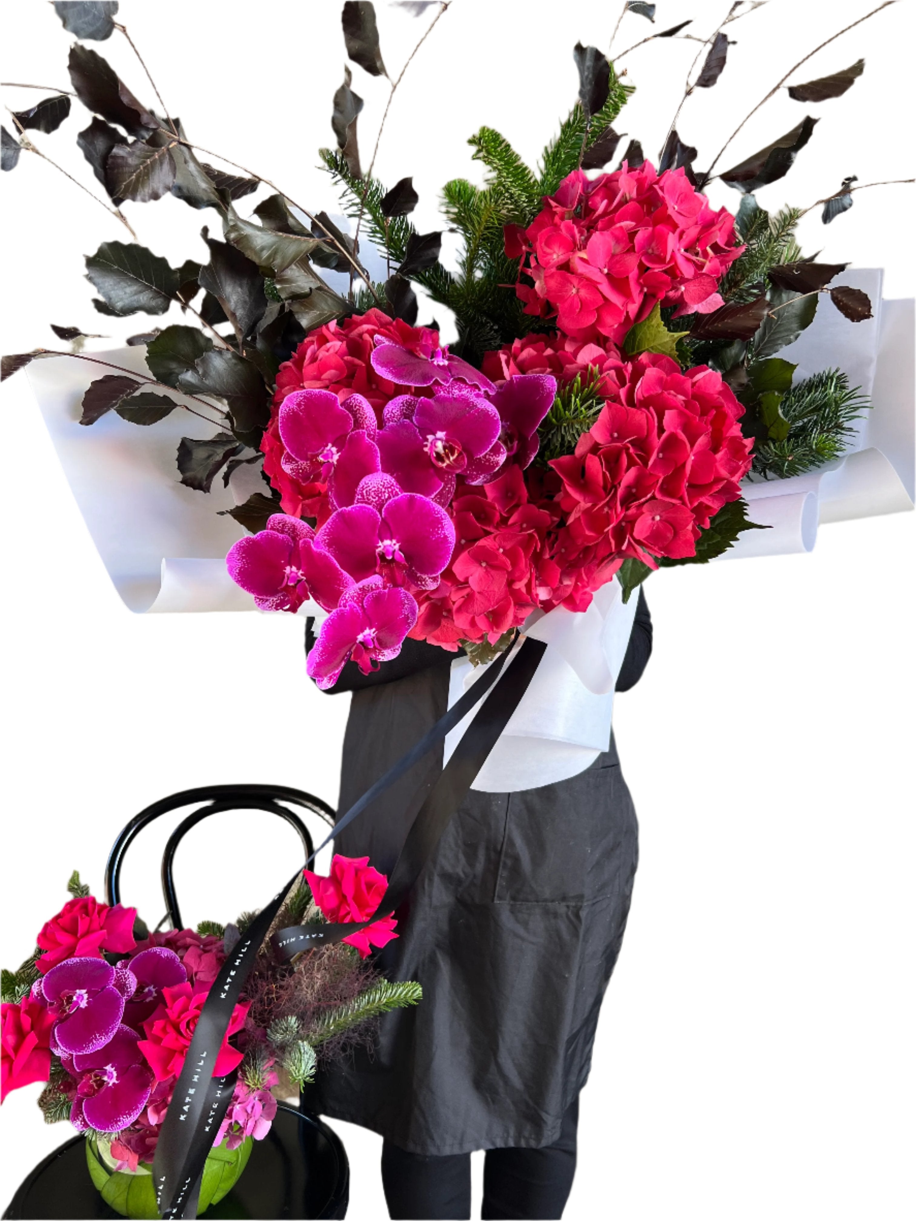 Florist holding a raspberry, burgundy and magenta Christmas flower bouquet and a complimentary vase design sitting on a lack bentwood chair with white wall background.