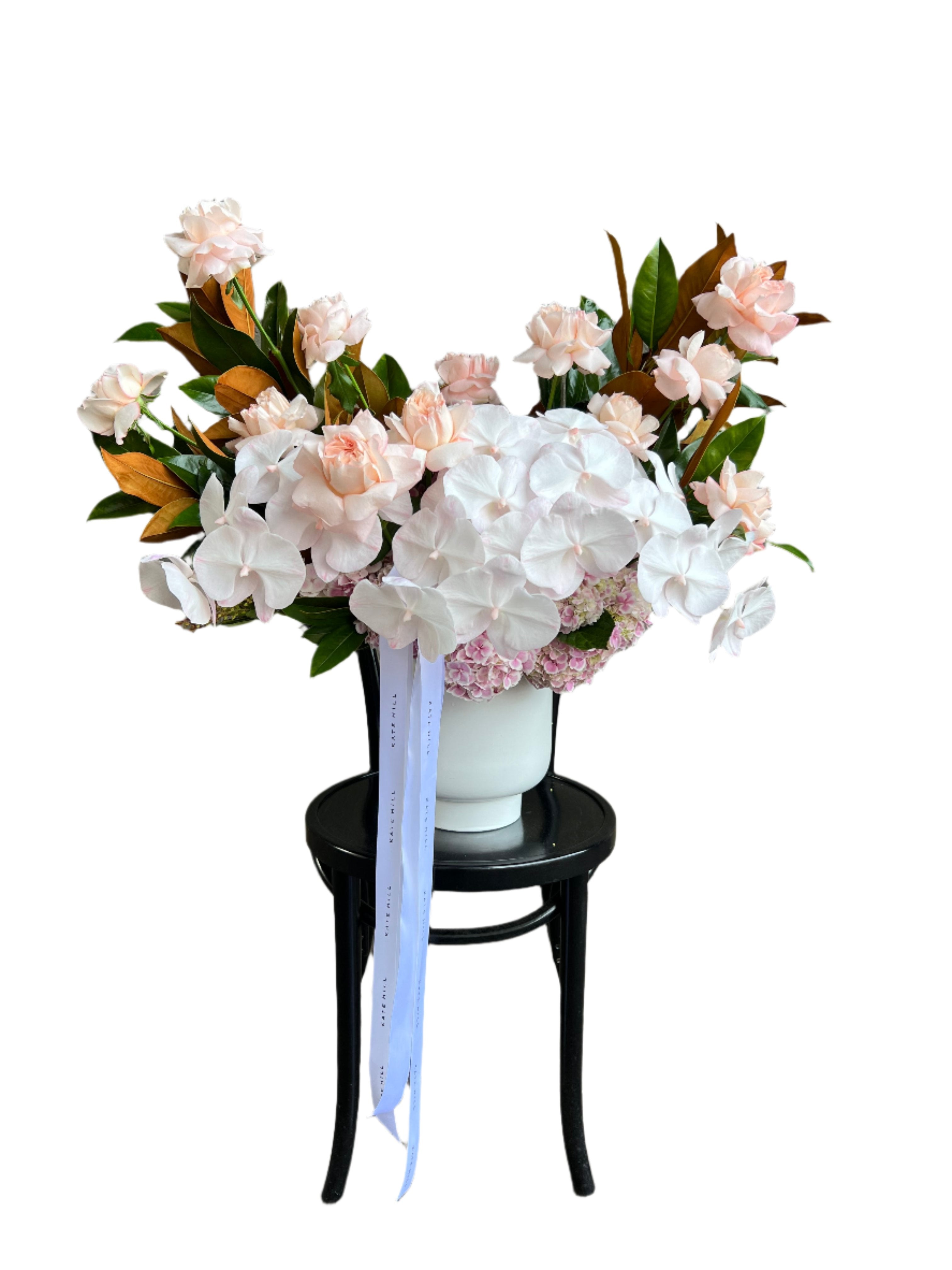 Large and luxe white ceramic vase displaying luxe blush pink flowers and magnolia foliage. Design sitting on a black bentwood chair with white wall behind.