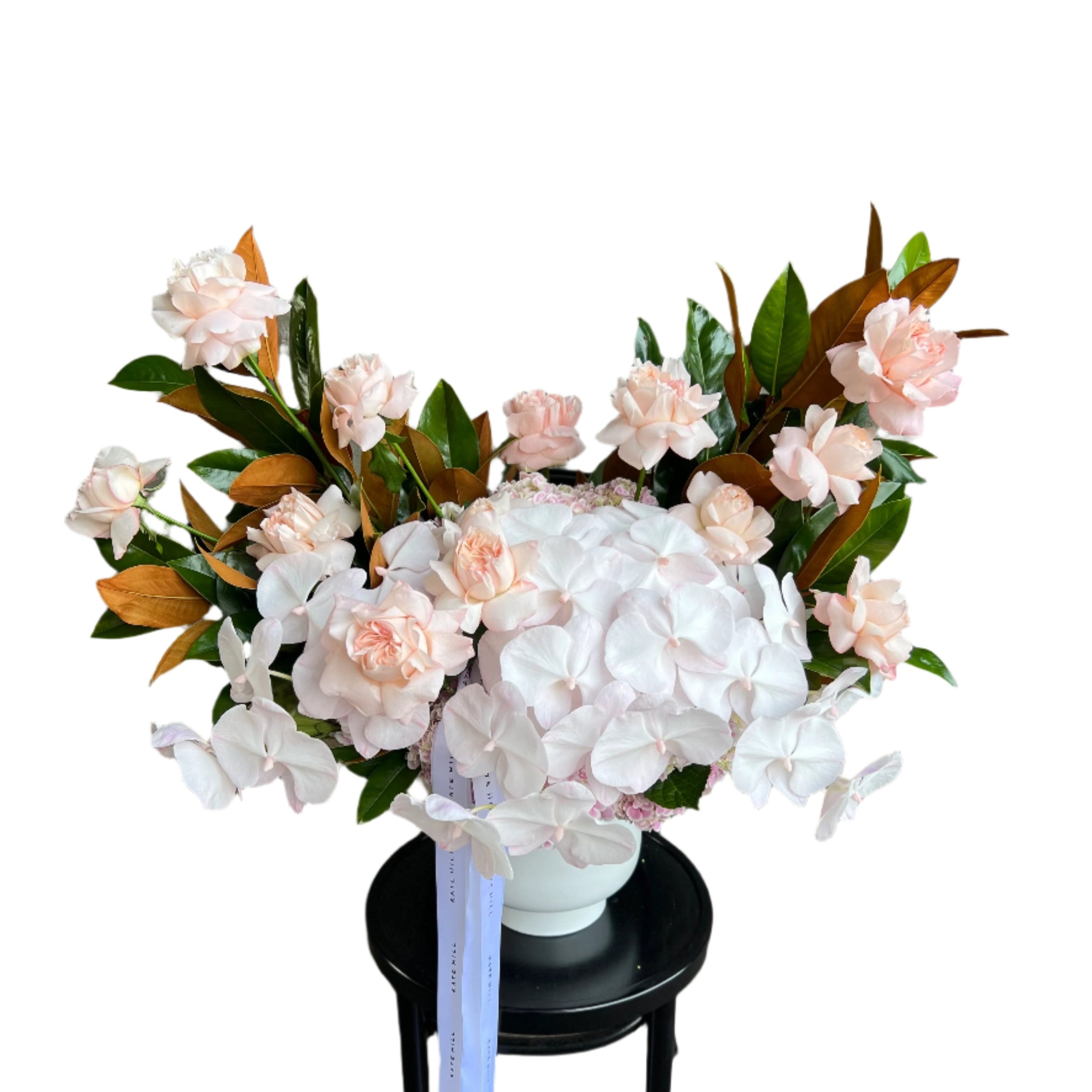 Large and luxe white ceramic vase displaying luxe blush pink flowers and magnolia foliage. Design sitting on a black bentwood chair with white wall behind.