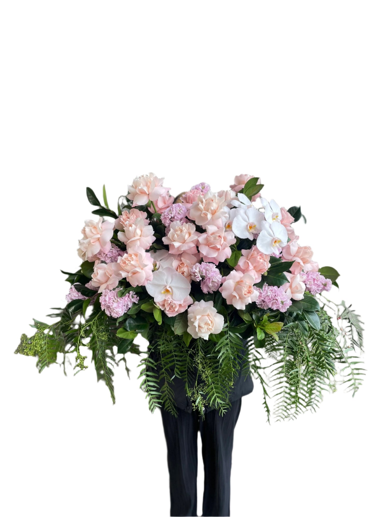Wide image of Classic blush roses, blush hydrangeas, lilac stock and foliages feature in a Pastel pink, white and green casket design, held by a florist wearing back, up against a white wall.