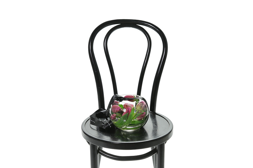 A 20cm glass ball vase, displaying pink tulip stems submerged inside the vase. Vase has black kate hill ribbon and is sitting on a black bentwood chair with a white background.
