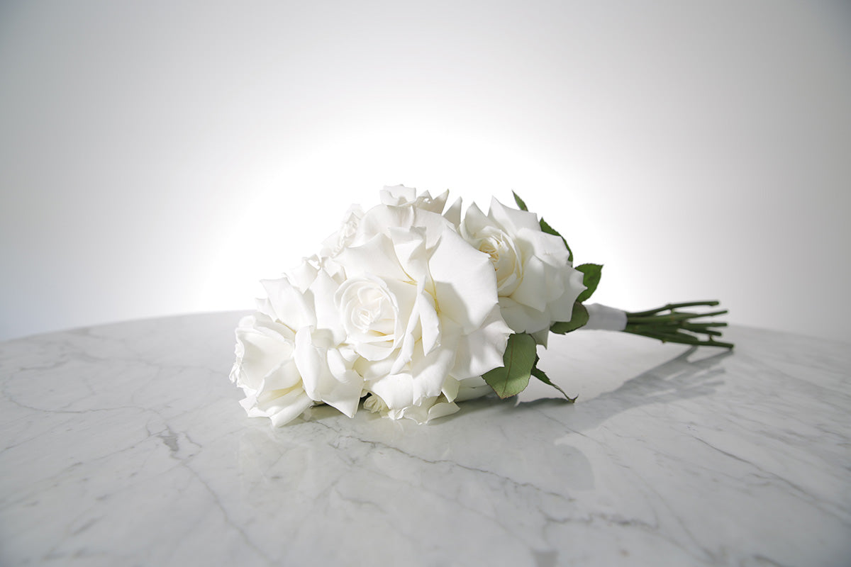 Reflexed white rose bridesmaid bouquet finished with ivory satin ribbon and pearl pin on a marble surface with a light grey background