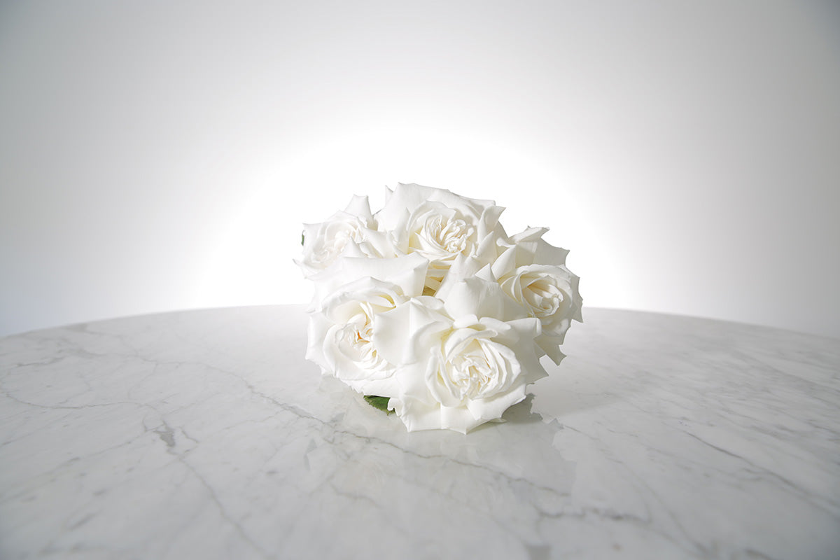 Reflexed white rose bridesmaid bouquet finished with ivory satin ribbon and pearl pin on a marble surface with a light grey background