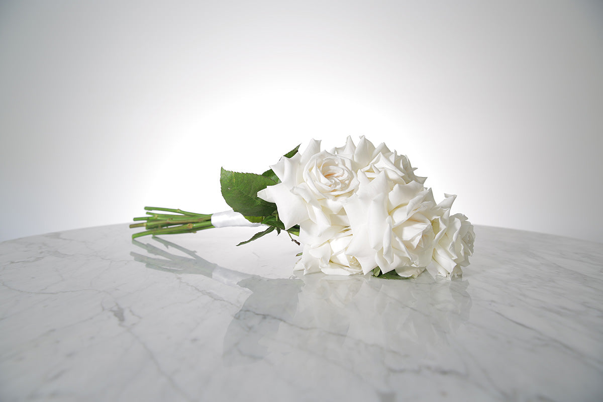 Reflexed white rose bridesmaid bouquet finished with ivory satin ribbon and pearl pin on a marble surface with a light grey background