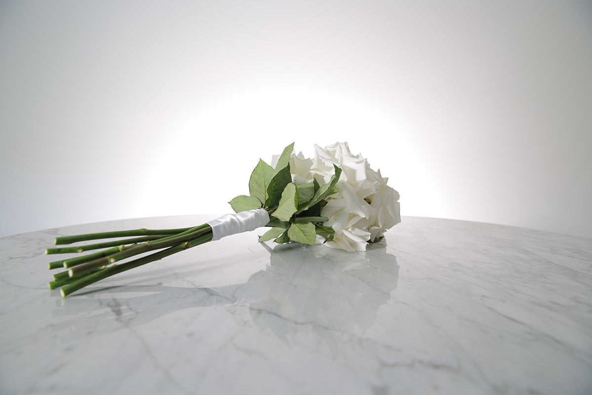 Reflexed white rose bridesmaid bouquet finished with ivory satin ribbon and pearl pin on a marble surface with a light grey background