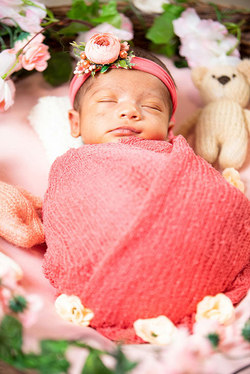 Newborn baby wrapped in pink fabric with a floral headband, surrounded by flowers and a teddy bear.