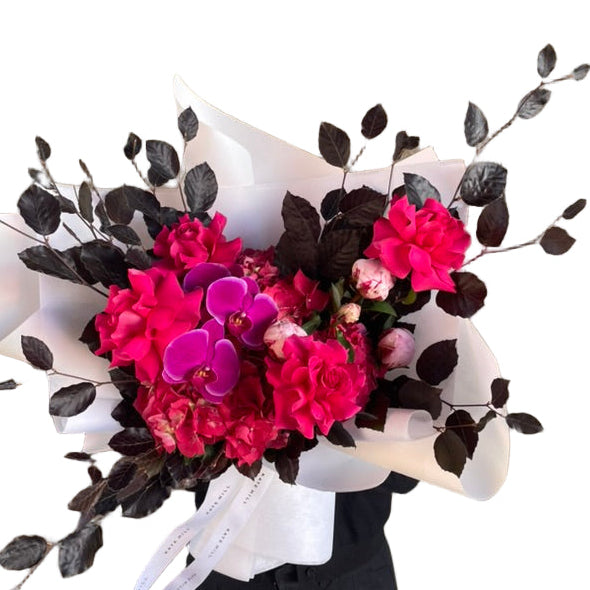 Close up image of a florist holding a large and bright bouquet in bright pink tones, wrapped beautifully in white and standing against a white wall.