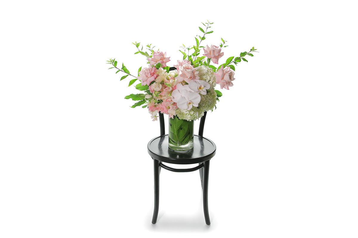 Wide image of Portia vase design. A 25cm cylinder tall vase design, featuring mixed soft pink and mid pink seasonal flowers. Tall cylinder vase design sitting on a black bentwood chair with white background. Flowers sitting on chair with white wall background.